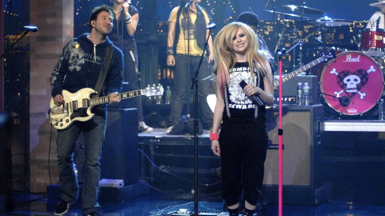 A band performs onstage, featuring a singer with long hair and a brightly colored outfit.
