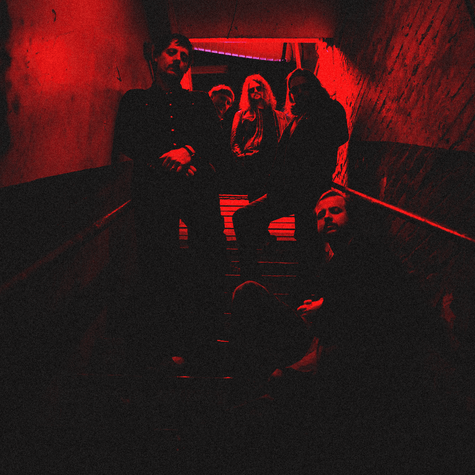A band poses dramatically on dimly lit stairs, illuminated with red lighting.