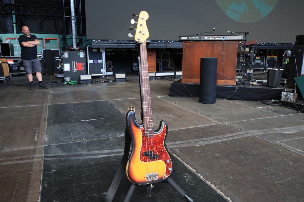 A bass guitar on a stand in a concert setting with a crew member in the background.