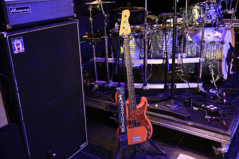 A bass guitar stands beside an amp and drum set on stage, with cables scattered around.