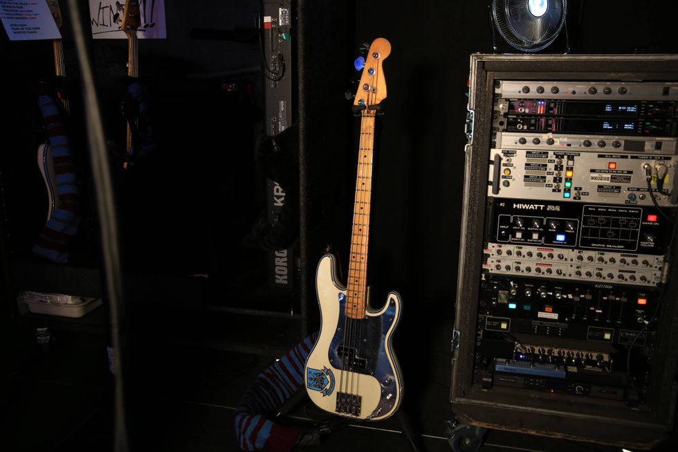 A bass guitar with a blue design stands beside a rack of music equipment.