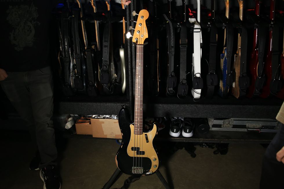 A black and gold bass guitar stands in front of a rack of various guitars.