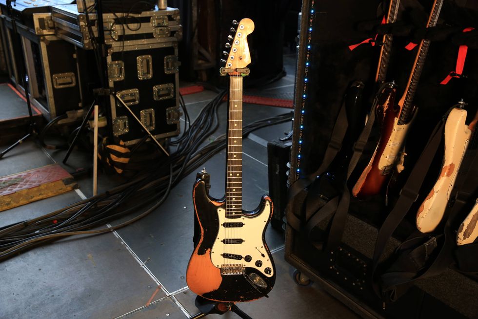 A black and tan electric guitar stands on stage next to guitar cases and cables.