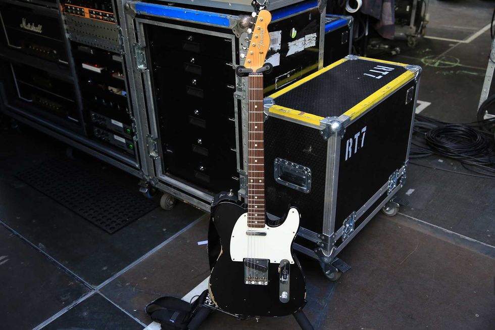 A black and white electric guitar rests on stage next to a road case and equipment.