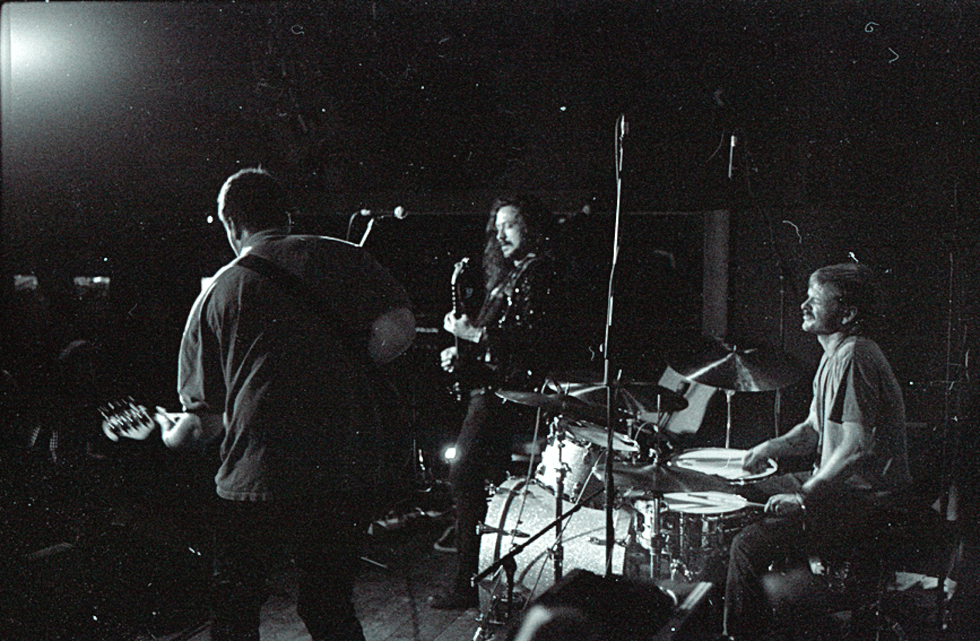 A black-and-white shot of a band performing on stage, featuring a guitarist and drummer.