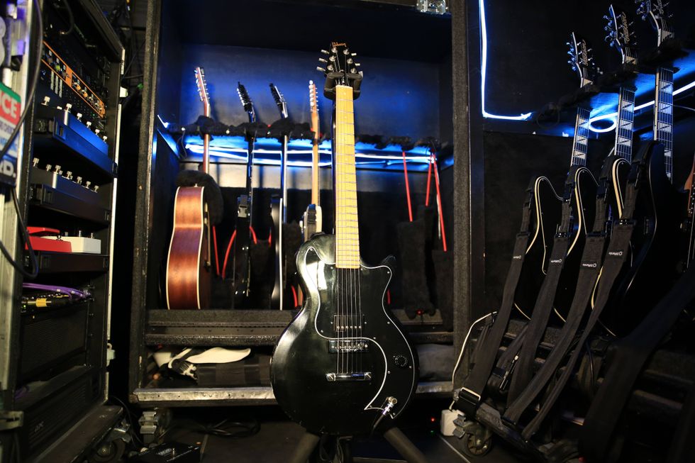 A black electric guitar stands in a music equipment rack filled with various instruments.