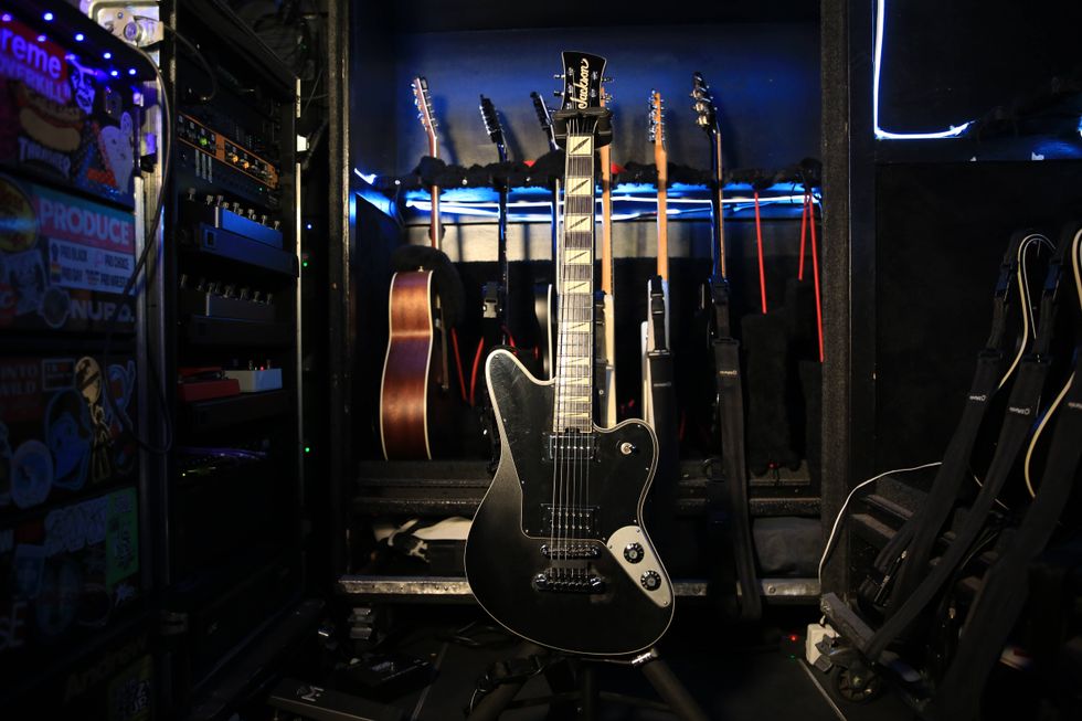 A black electric guitar stands in front of a collection of other guitars in a case.