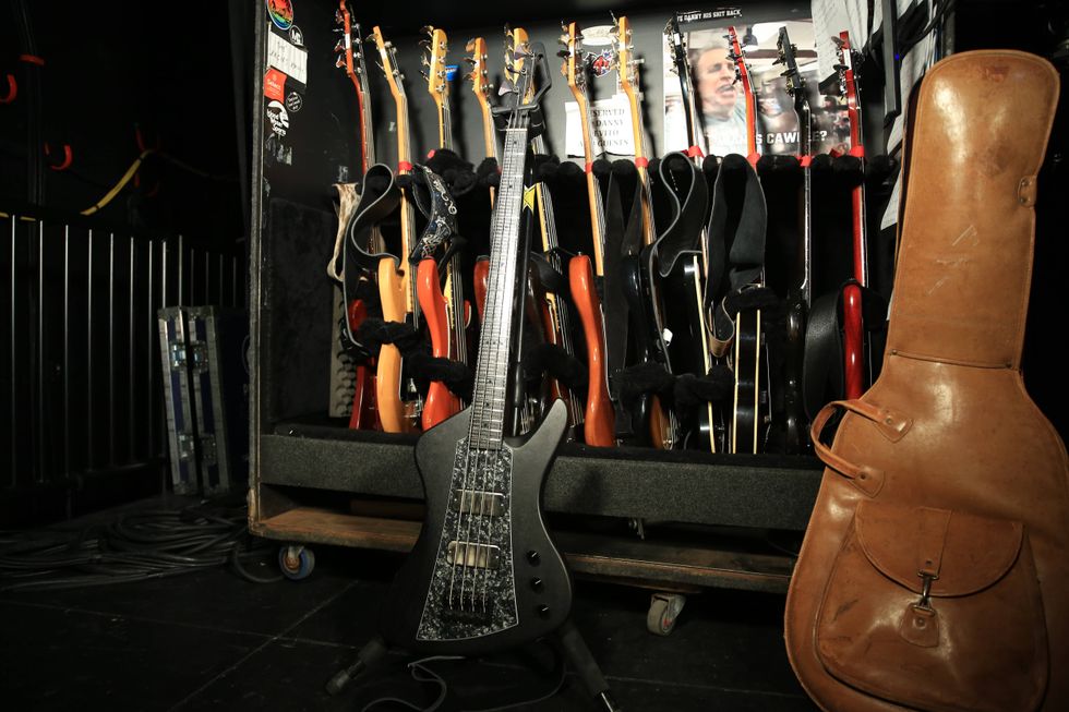 A black electric guitar stands in front of a rack filled with various guitars and a case.