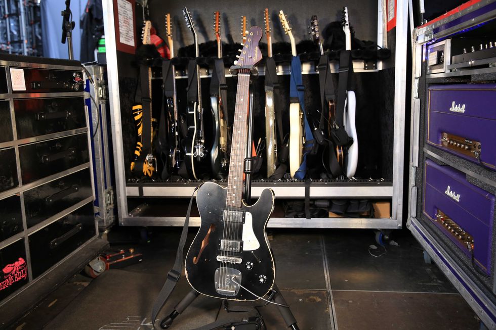 A black electric guitar stands in front of a rack filled with various other guitars.