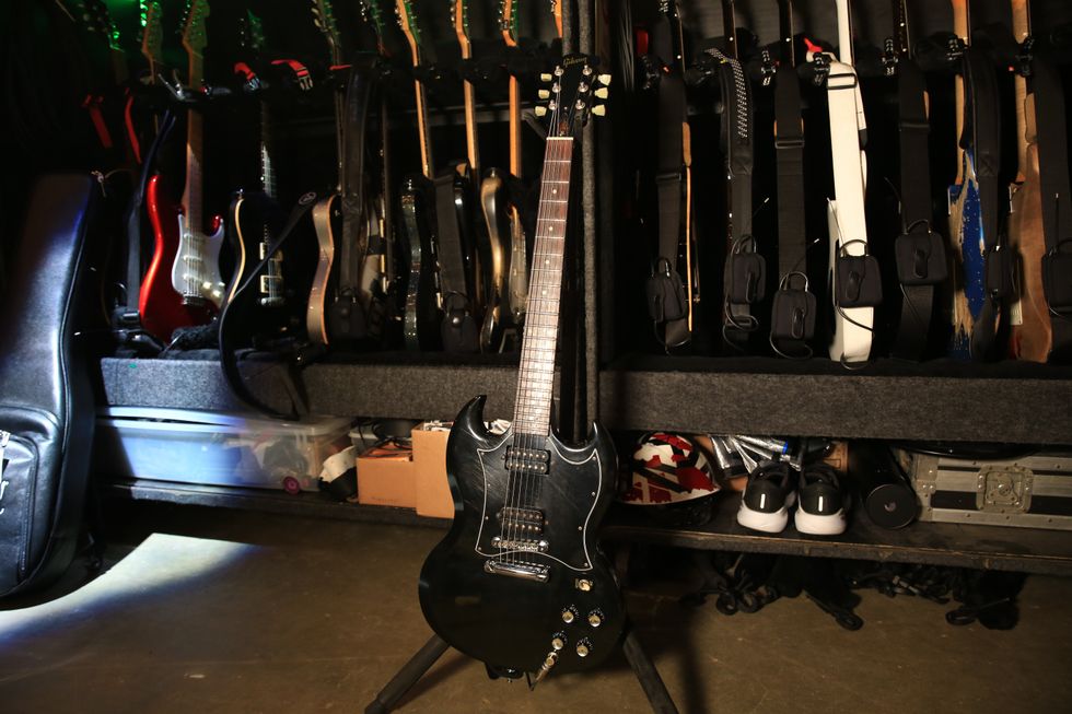 A black electric guitar stands in front of a wall of various guitars and accessories.