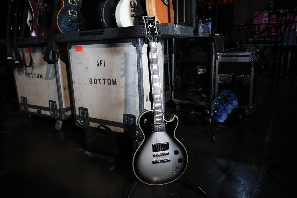A black electric guitar stands in front of equipment cases at a concert venue.