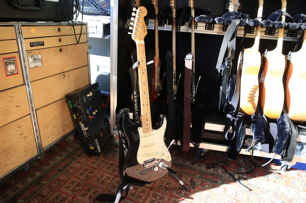 A black electric guitar stands next to a wooden cabinet and additional guitars on a rack.