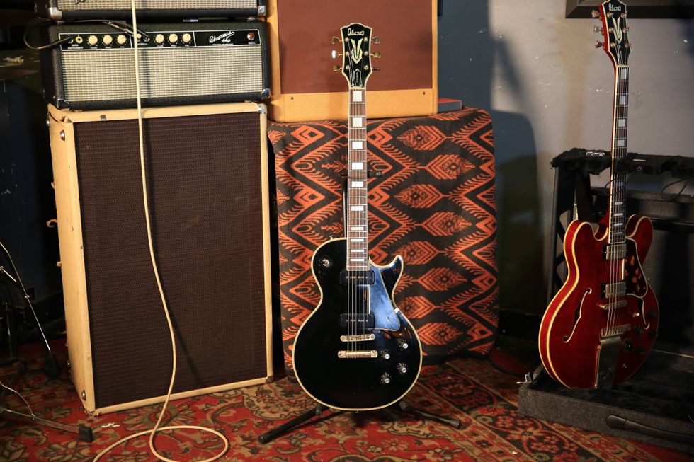 A black electric guitar stands next to amplifiers on a patterned rug.