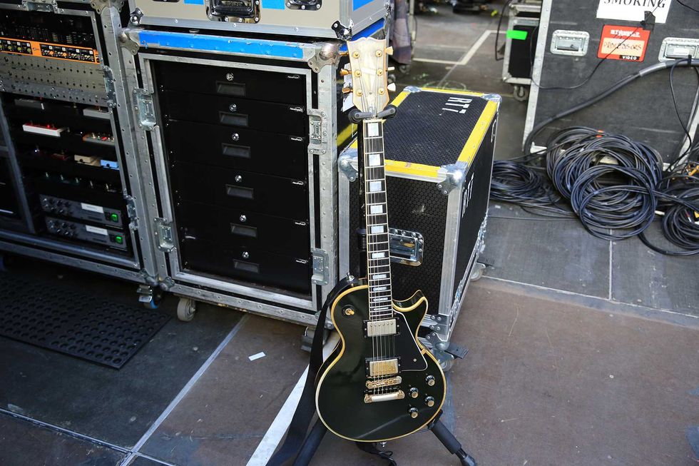 A black electric guitar stands next to sound equipment and tangled cables on stage.