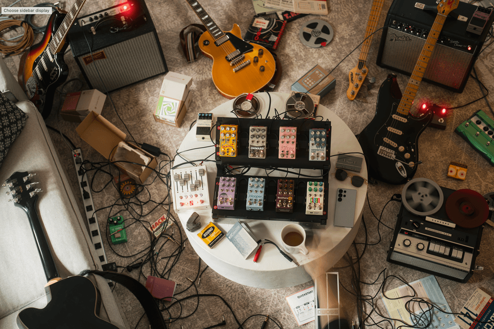 A cluttered workspace featuring guitars, pedals, and recording equipment on a coffee table.