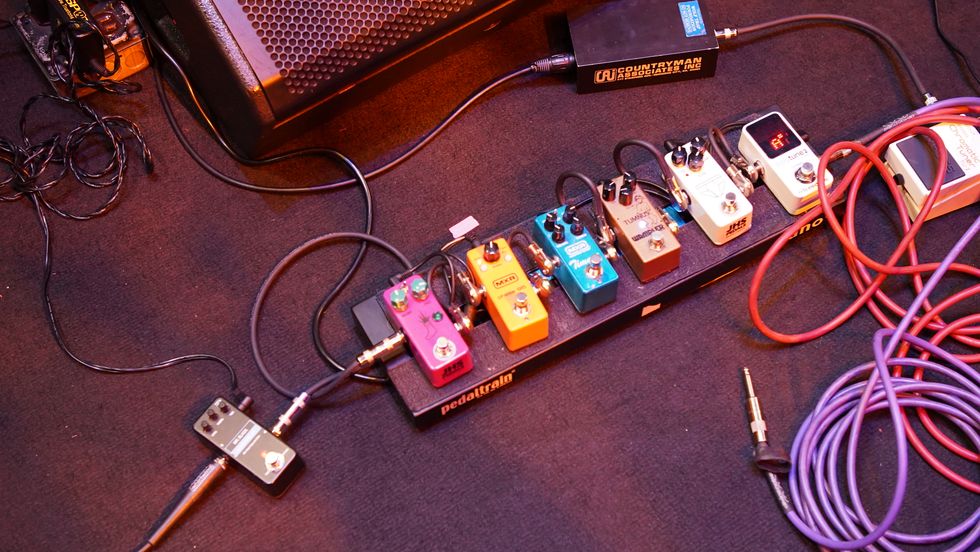 A collection of guitar pedals arranged on a board with cables and an amplifier in the background.