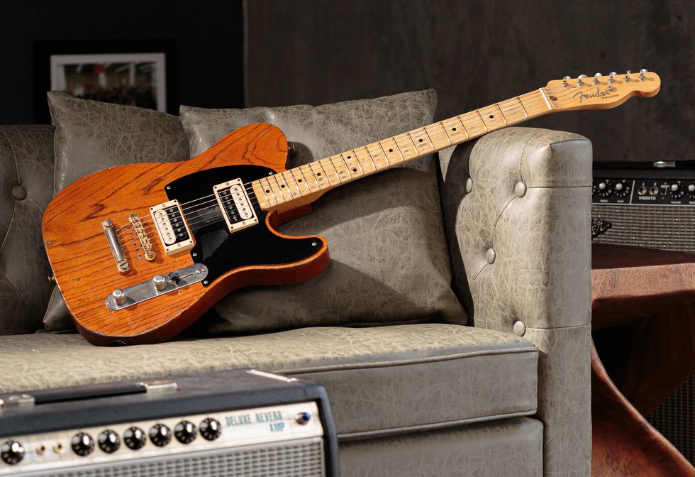 A Fender Telecaster guitar rests on a stylish gray couch, with amplifiers in the background.