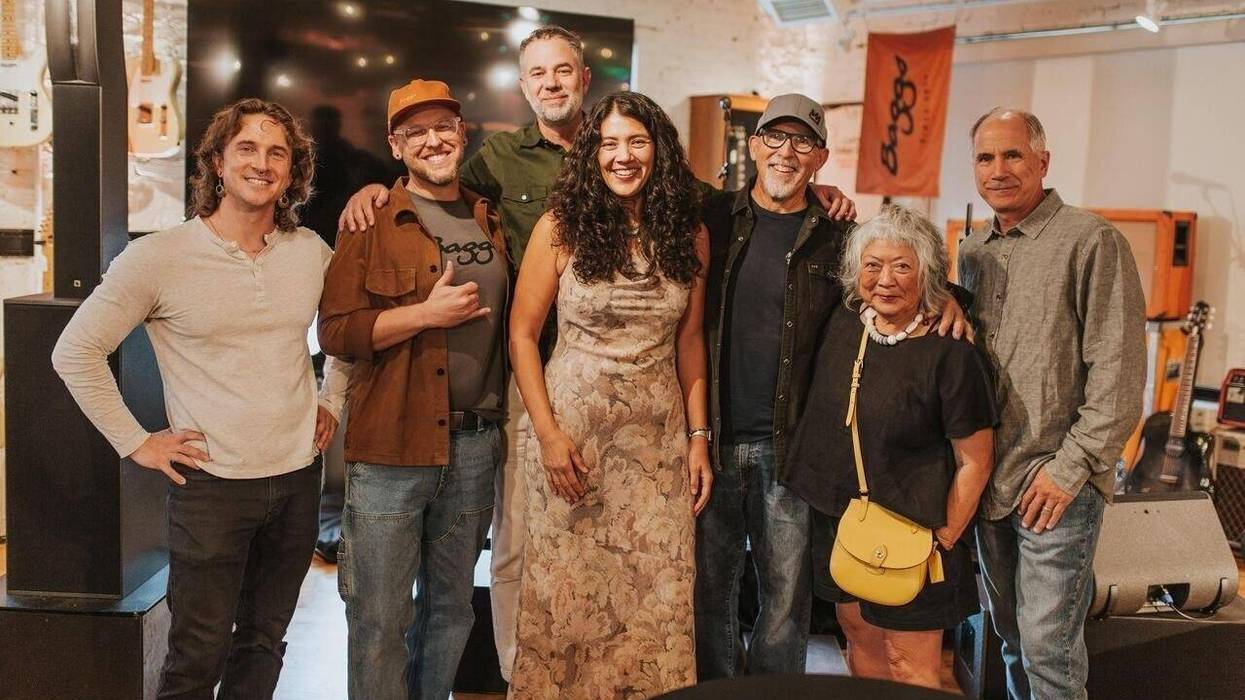 A group of seven people pose together in a music store, smiling and dressed casually.