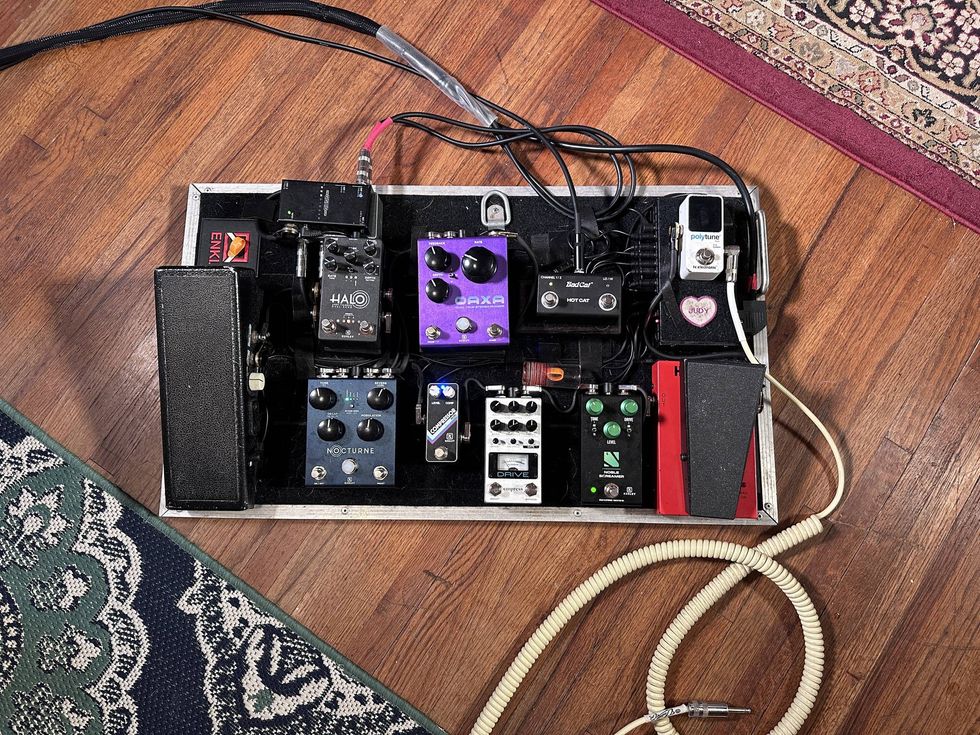 A guitar pedalboard with various effects pedals and connected cables on a wooden floor.