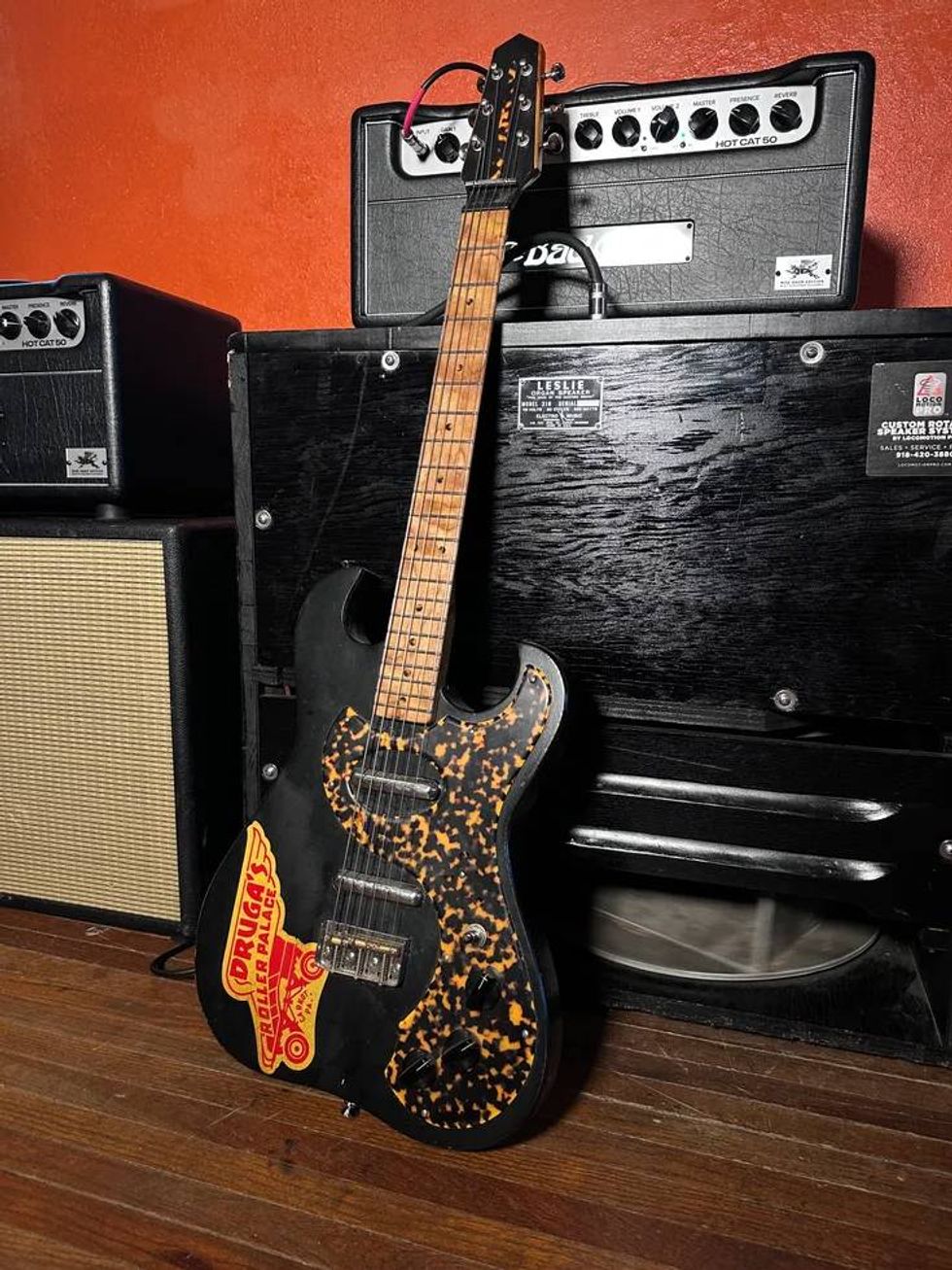 A guitar with a tortoiseshell finish, leaning against amplifiers on a wooden floor.