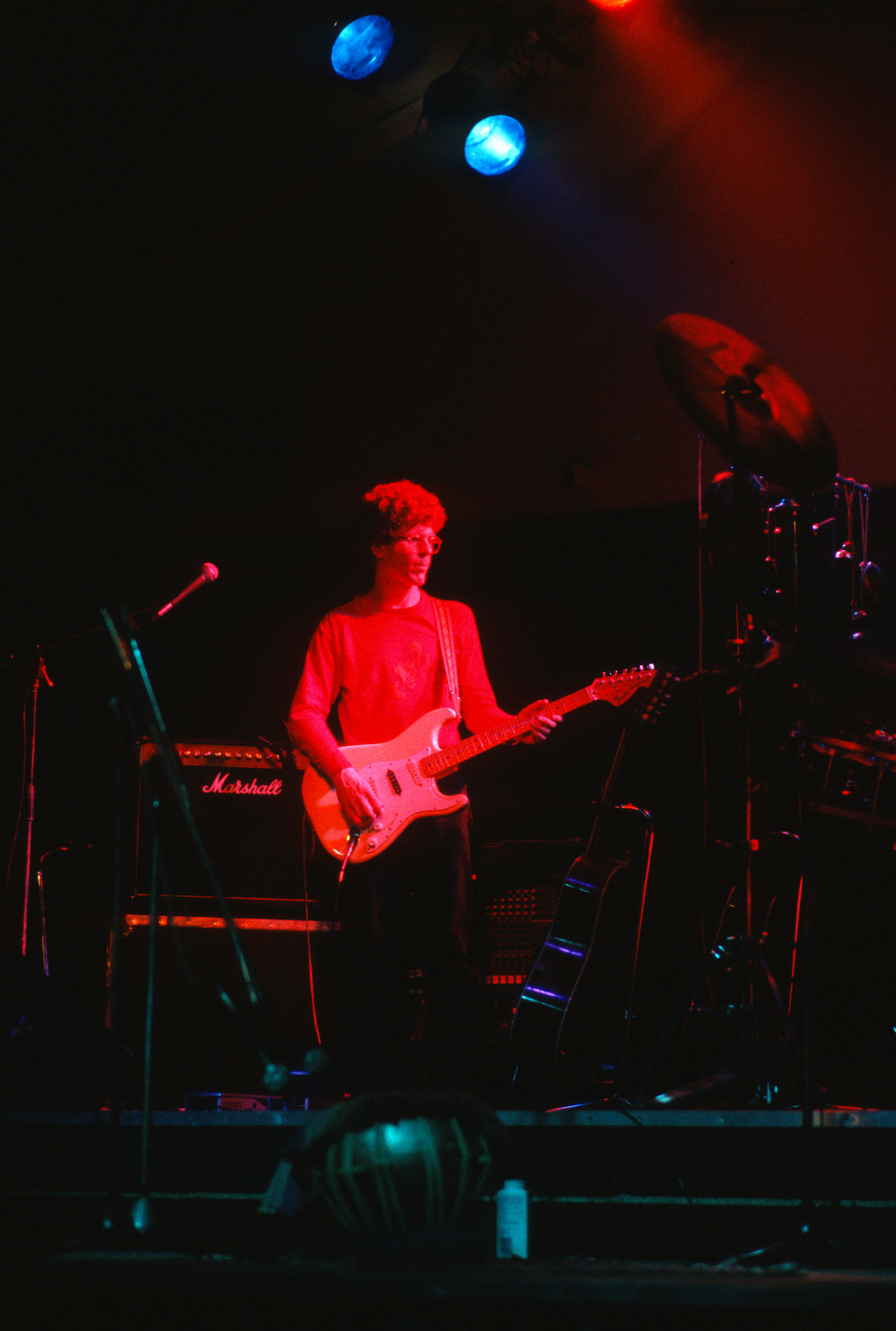 A guitarist in a red shirt performs on stage with vibrant lighting and musical equipment around.