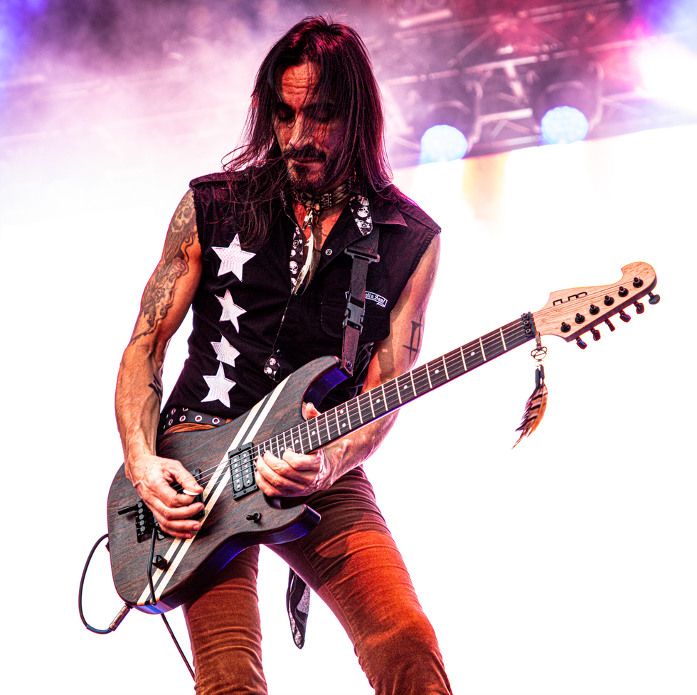 A guitarist with long hair performs on stage, wearing a sleeveless shirt with stars.