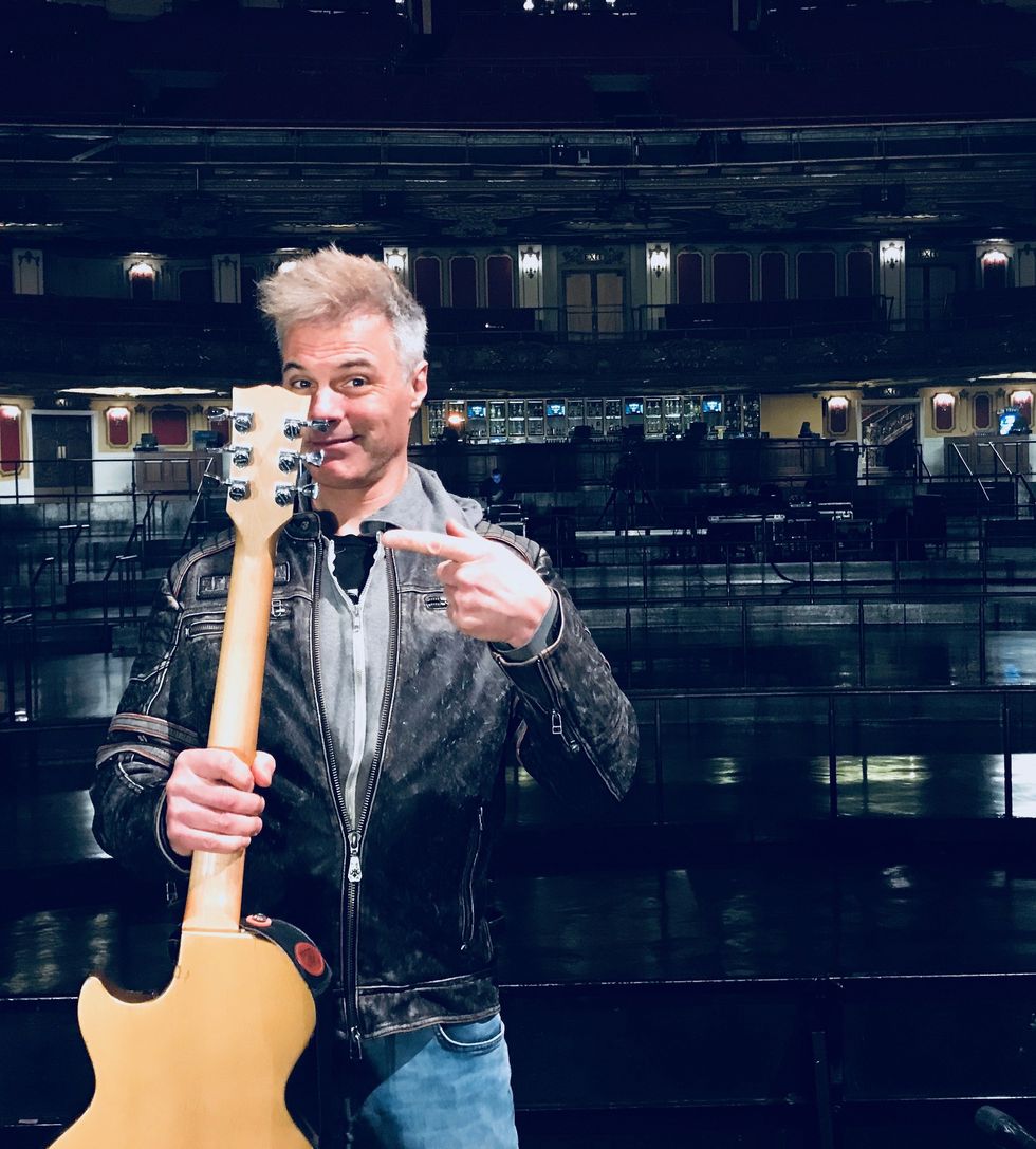 A man in a leather jacket points at his guitar backstage in a concert venue.