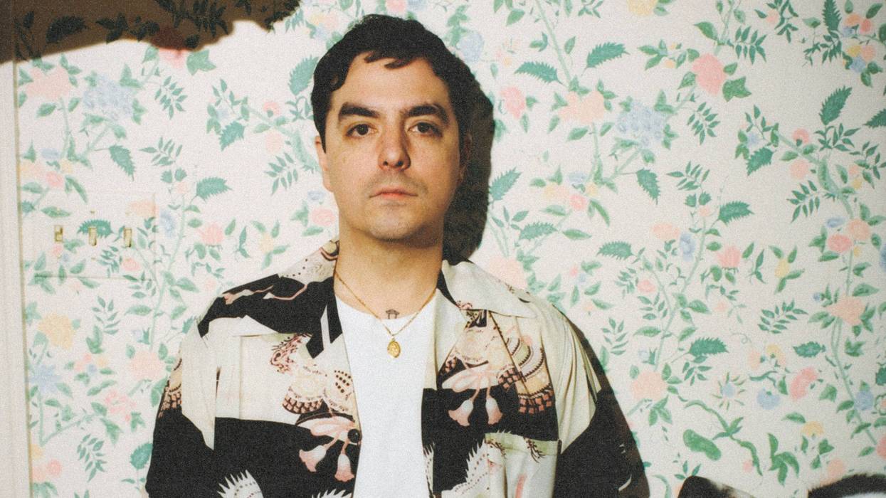 A man in a patterned shirt sits next to a dog, surrounded by floral wallpaper.