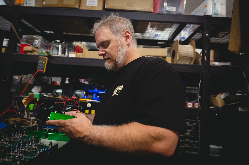 A man inspects a green electronic component in a cluttered workshop.