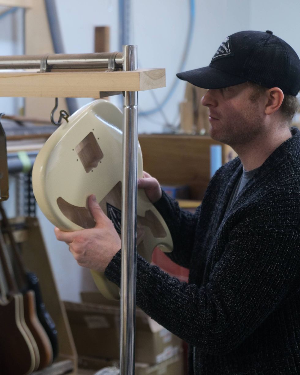 A man inspects a guitar body on a rack in a workshop.