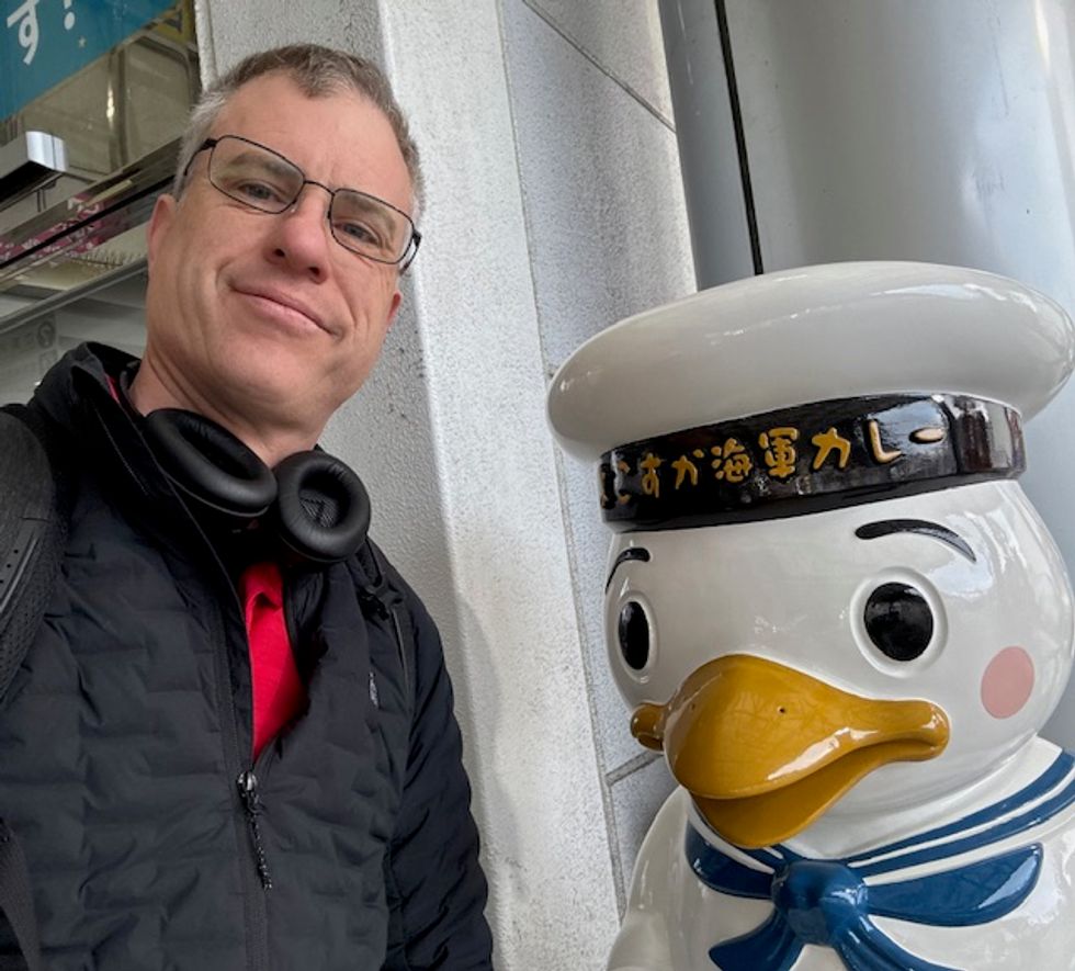 A man poses next to a whimsical sailor duck statue in an urban setting.