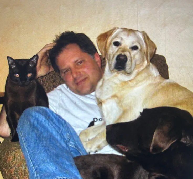 A man sits on a couch surrounded by a cat and two dogs, smiling contentedly.