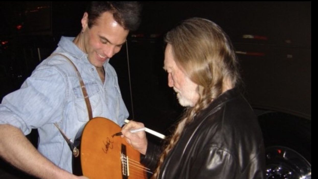 A man smiles as another signs an acoustic guitar in a backstage setting.