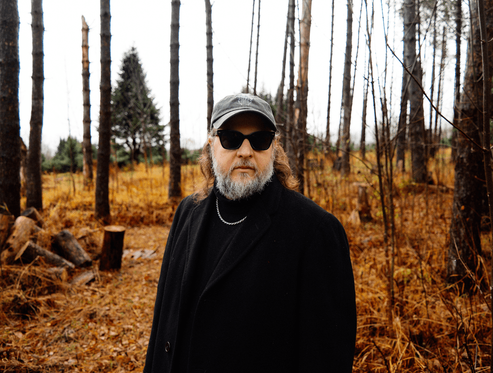 A man with glasses and a cap stands amidst tall trees in a forest during fall.
