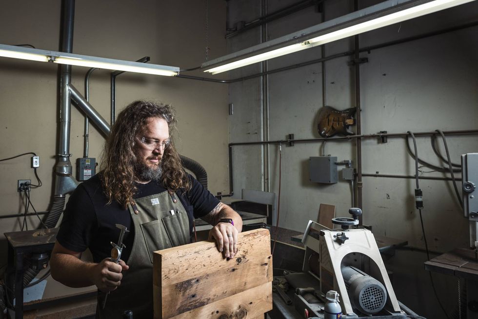 A man with long hair and glasses works with wood in a workshop, holding a chisel.