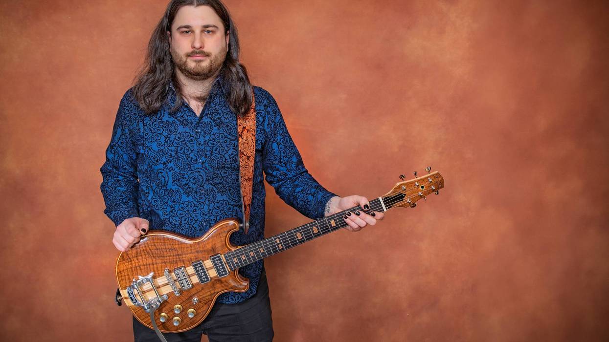 A man with long hair in a blue patterned shirt holds a unique guitar against a brown backdrop.
