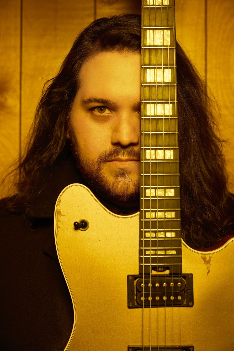 A man with long hair partially hides his face behind a vintage gold guitar.