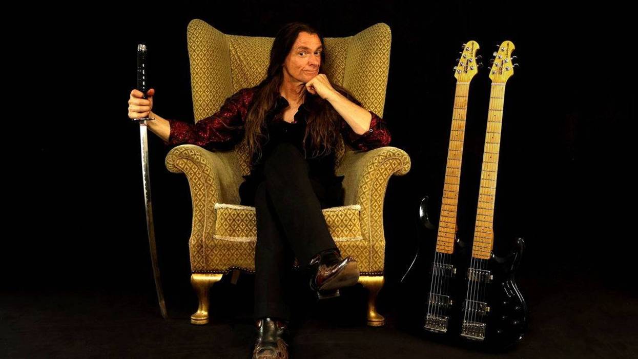 A man with long hair sits in a chair, holding a sword, beside two electric guitars.