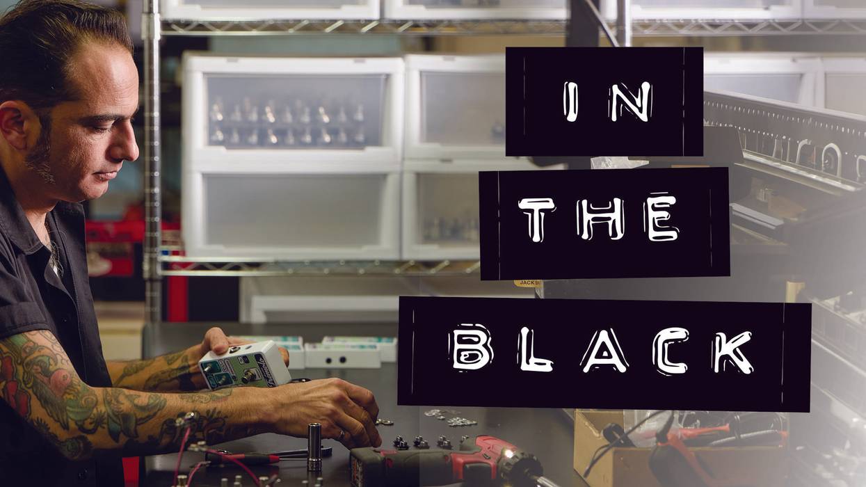 A man with tattoos works on electronic equipment in a workshop, surrounded by tools and parts.