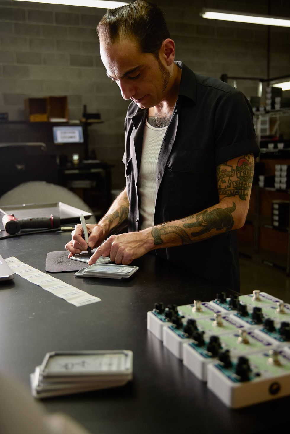 A man with tattoos writes on a device at a workbench surrounded by equipment and labels.