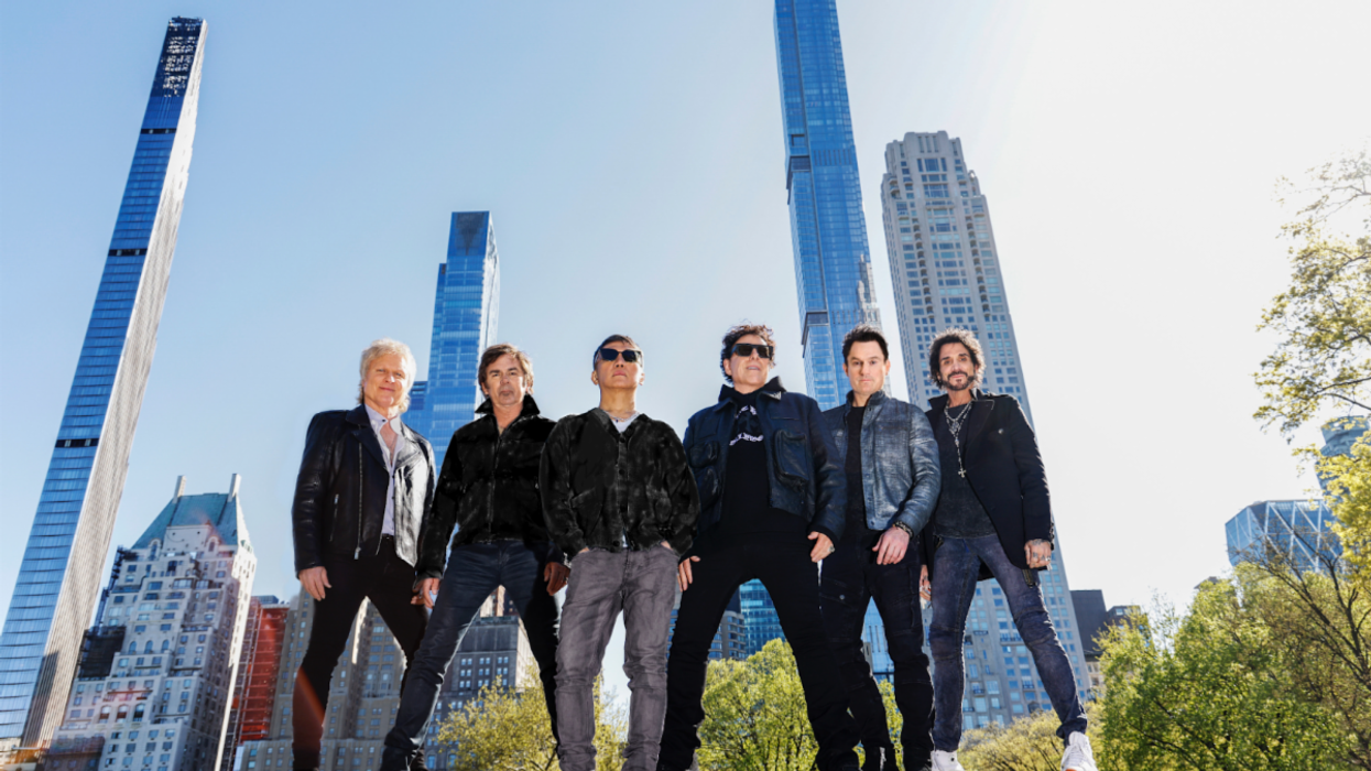 A music group poses confidently in front of tall skyscrapers on a clear day.