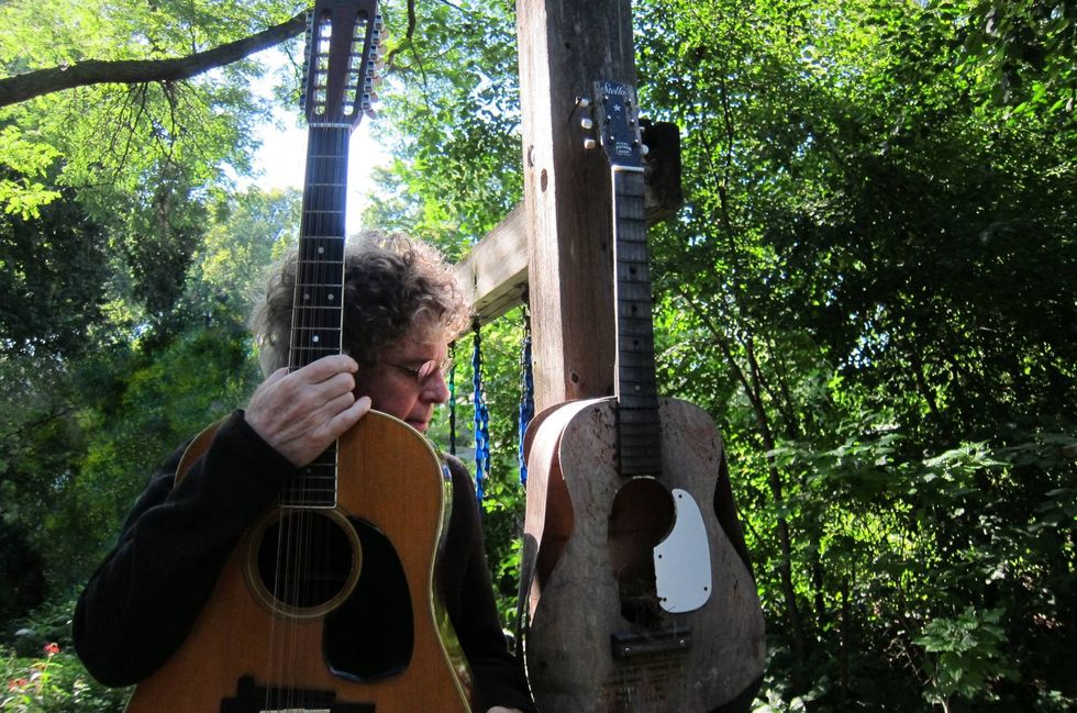 A musician holding an acoustic guitar beside an old, hanging guitar, surrounded by greenery.