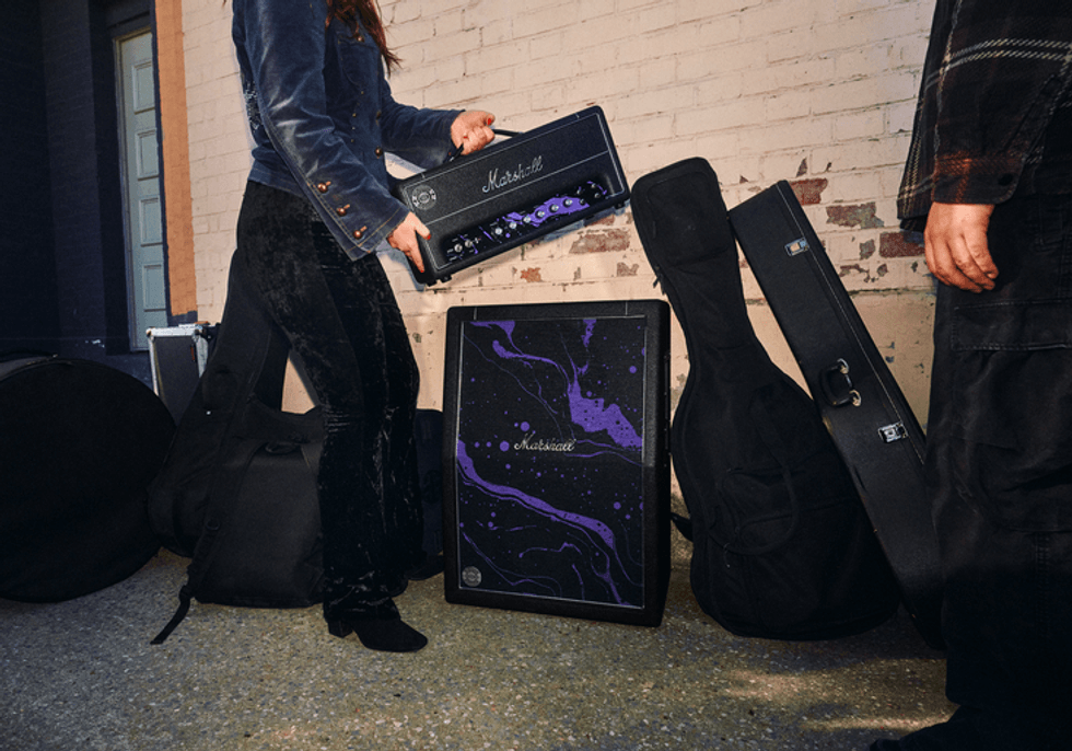 A musician lifts a Marshall amp, surrounded by guitars and gear on the ground.