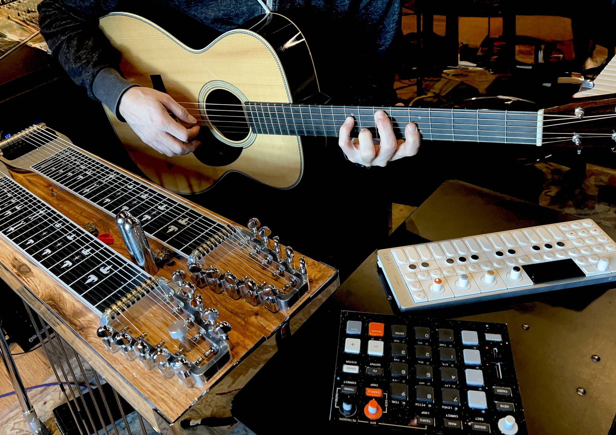 A musician plays an acoustic guitar alongside a lap steel guitar and digital music devices.
