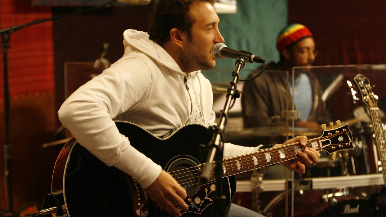 A musician plays an acoustic guitar and sings in a studio, while a drummer accompanies him.