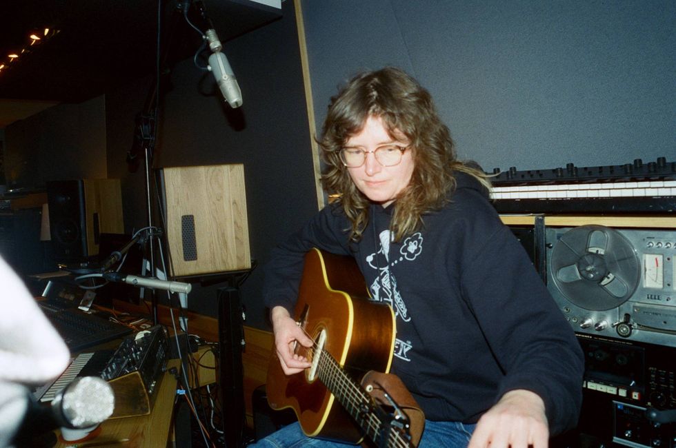 A musician plays an acoustic guitar in a recording studio, surrounded by audio equipment.