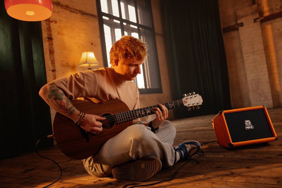 A musician sits on the floor playing guitar beside an orange amplifier in a cozy room.