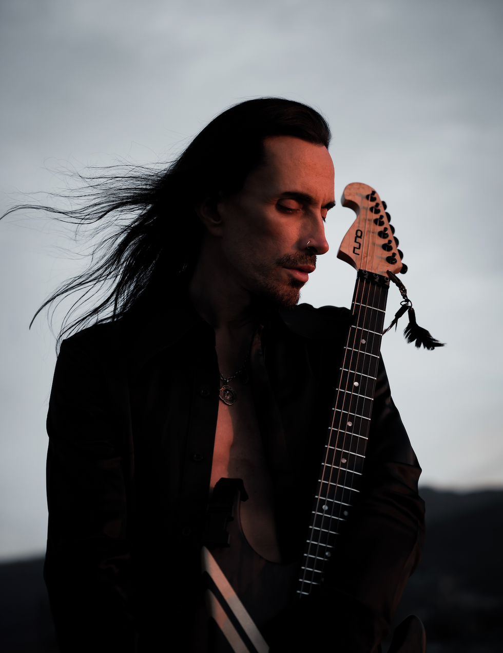 A musician with long hair holds an electric guitar against a dramatic sunset backdrop.