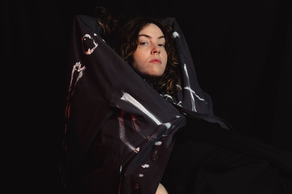 A person with curly hair poses dramatically in a dark, painted coat against a black background.