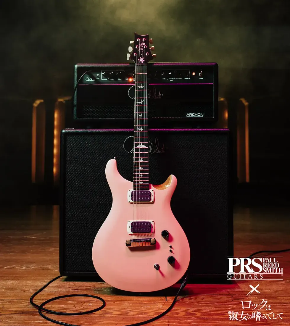 A pink electric guitar stands in front of a black amplifier on a wooden stage.